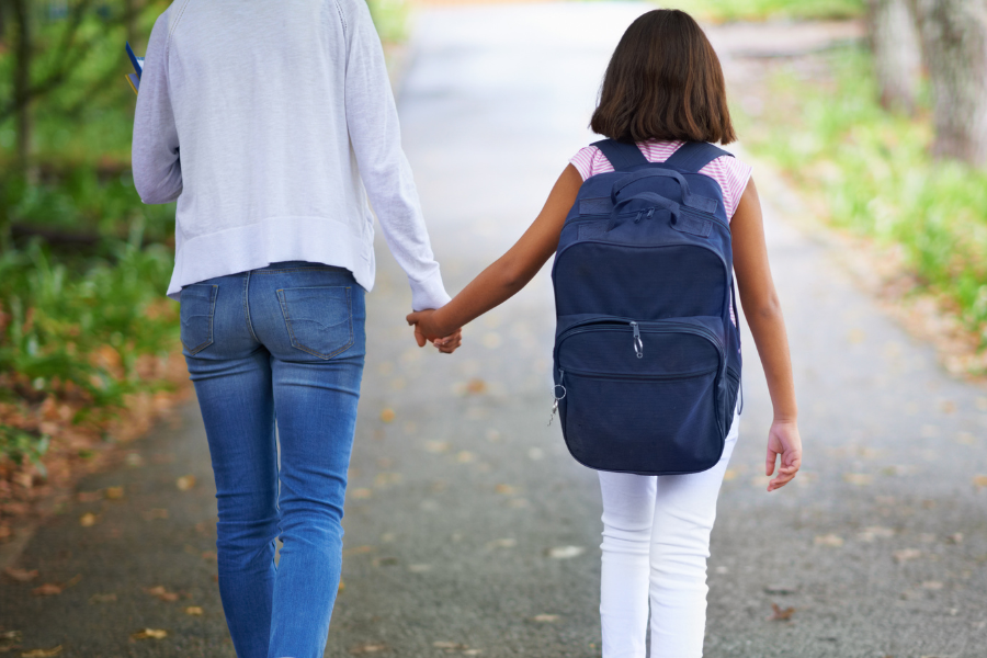 Mother and school-aged child with backpack walk down path to school.